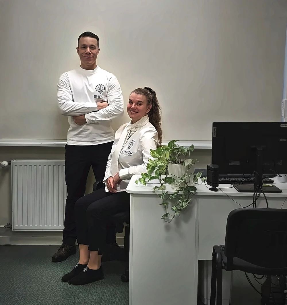 The two founders of Empor Solutions in white Empor shirts in their first office, one standing with crossed arms, the other seated at the desk, both smiling.
